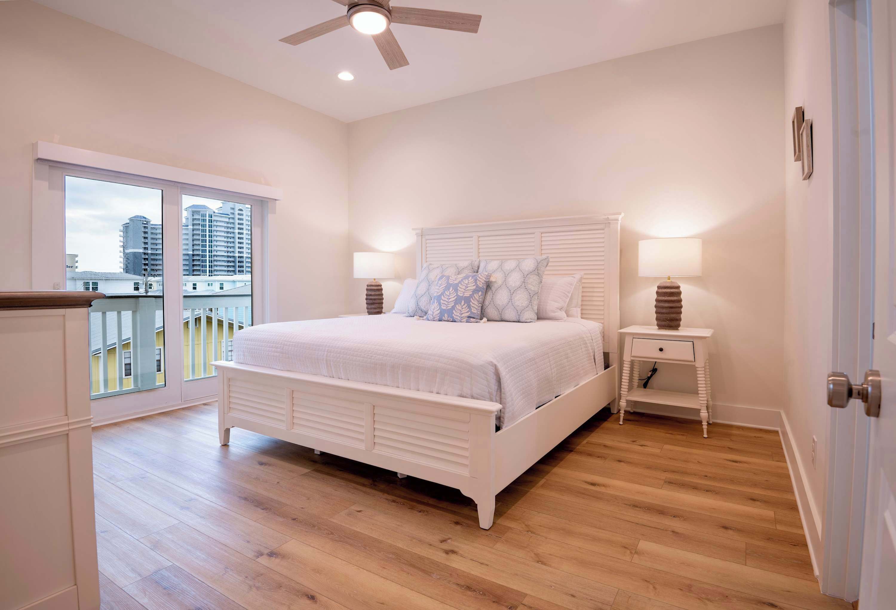A bedroom with a large bed, two bedside tables with lamps, a ceiling fan, and a balcony with a view of buildings.
