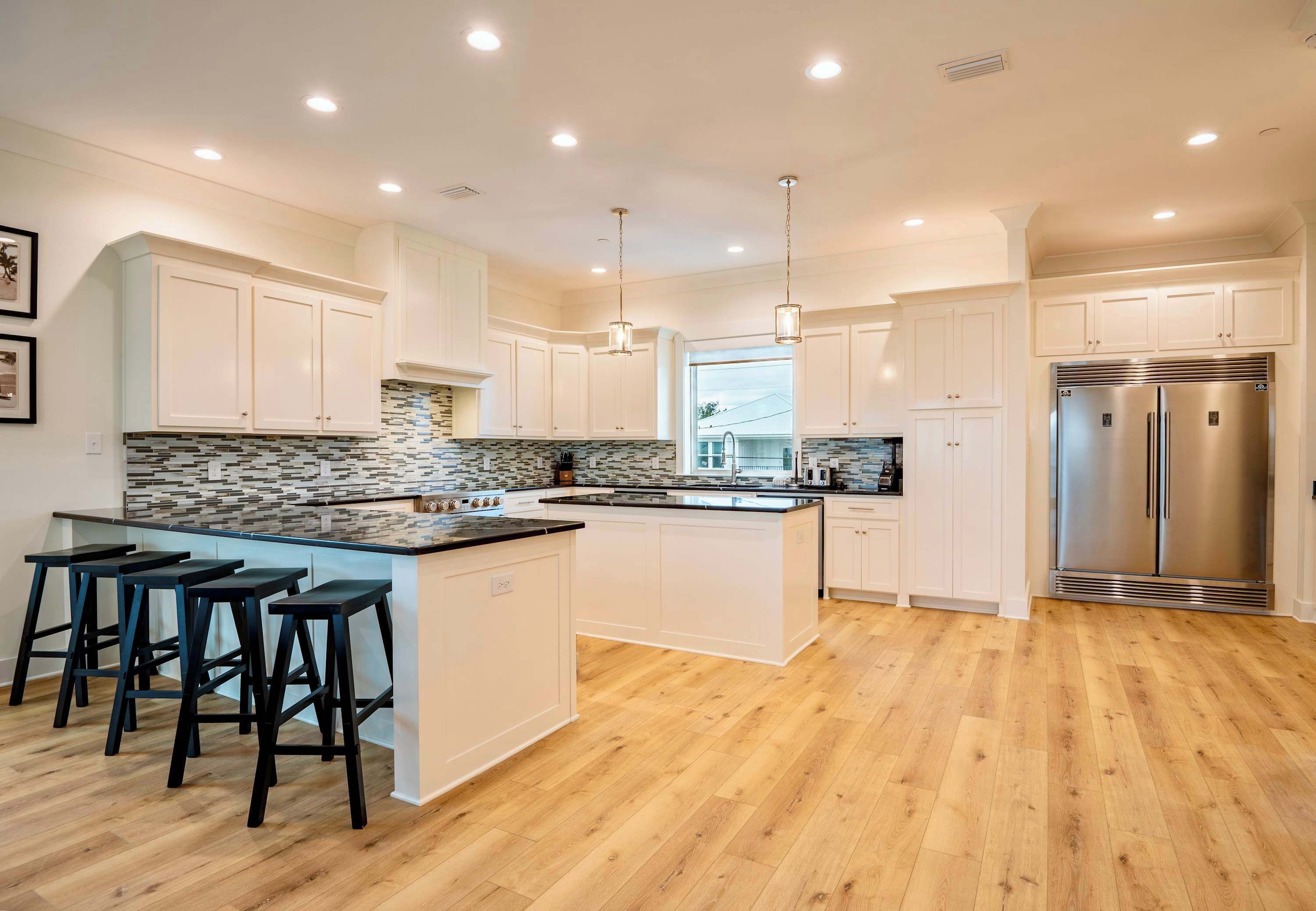A modern kitchen with white cabinetry, a large island with bar stools, a stainless steel refrigerator, and a patterned backsplash.