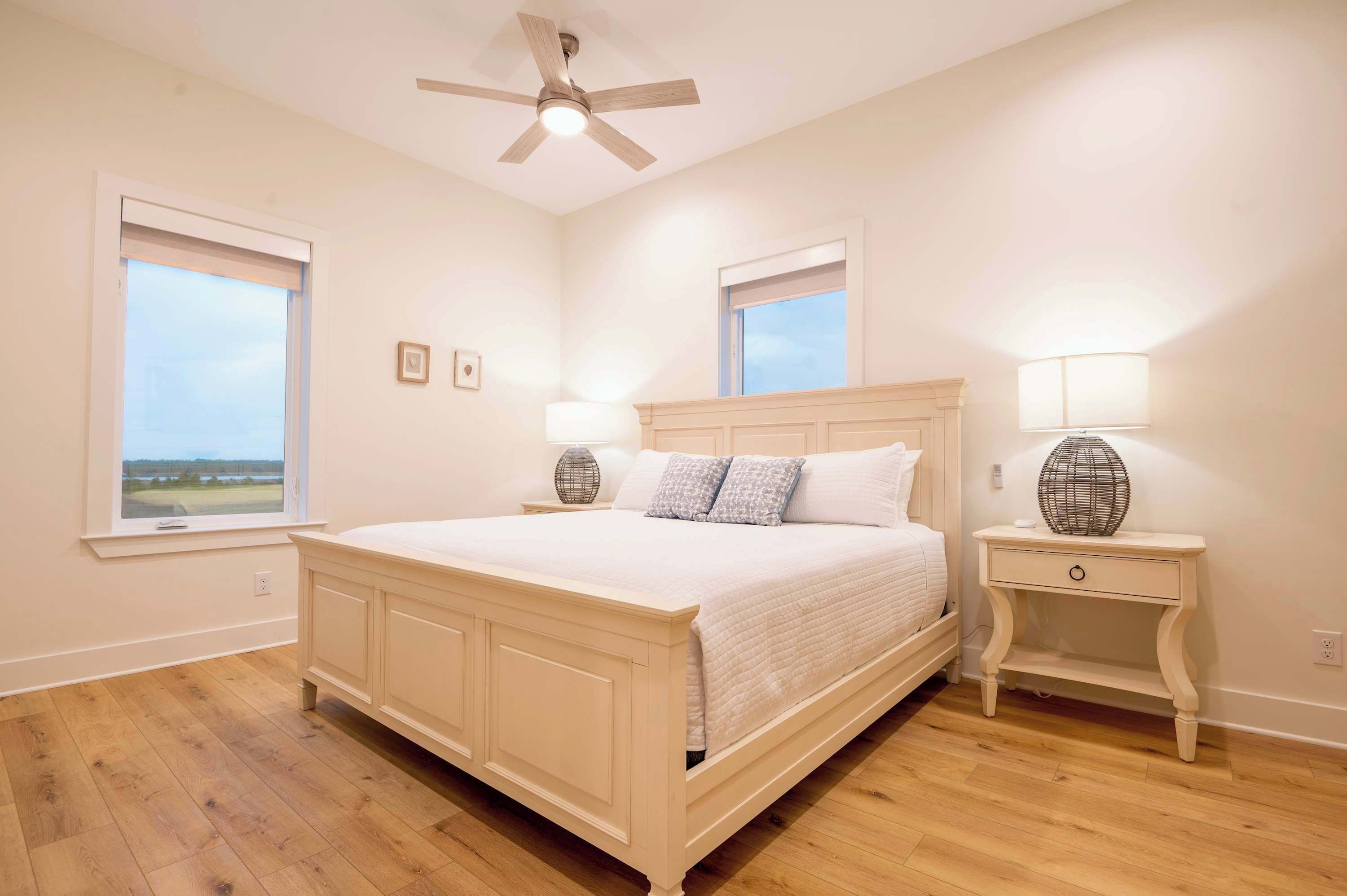 Un dormitorio con una cama grande, dos mesitas de noche con lámparas, un ventilador de techo y ventanas con persianas.