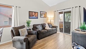 Een woonkamer met een bruine leren bank, twee fauteuils, een bijzettafeltje met een plant en een lamp. Twee ingelijste foto's hangen aan de muur en een schuifdeur leidt naar een terras.
