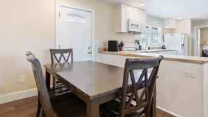 A modern kitchen with white cabinets, a microwave, a refrigerator, and a dining area with a wooden table and chairs. - Diana's Place (Murfreesboro)