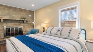 A bedroom with a stone fireplace, a bed with striped bedding, a blue chair, and a window with blinds. - Diana's Place (Murfreesboro)