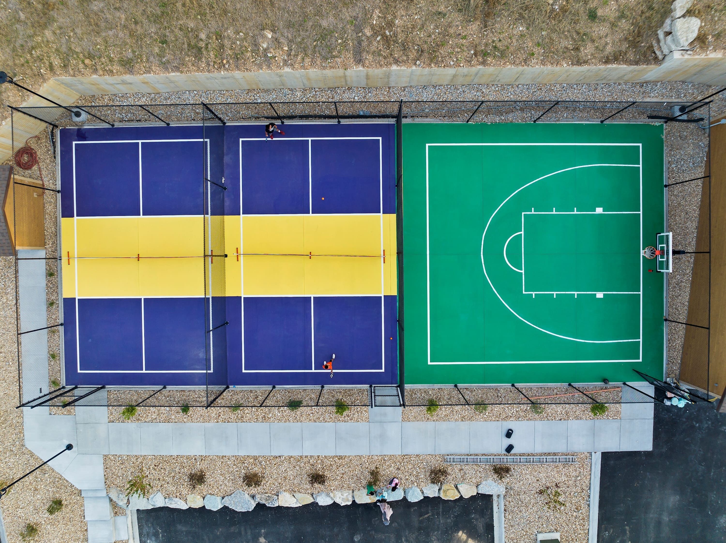 Vista aérea de uma quadra poliesportiva com campos de basquete e tênis, cercada por uma grade e um caminho de cascalho.