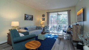 Una sala de estar acogedora con un sofá, un sillón y una mesita redonda. Hay un balcón con vistas a árboles.
