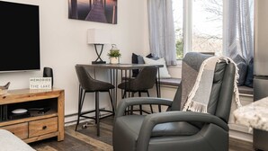 Un soggiorno moderno con una grande TV a schermo piatto, un tavolino in legno con un altoparlante, una zona giorno accogliente con una panca e poltrone, e una finestra con tende.