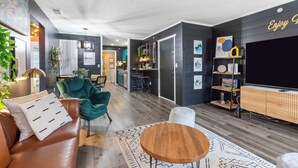 Ein modernes Wohnzimmer mit einer Ledersofa, einem grünen Sessel und einem Holztisch. Im Hintergrund ist eine Küchenzeile mit einer Bar und einer Neonreklame zu sehen.