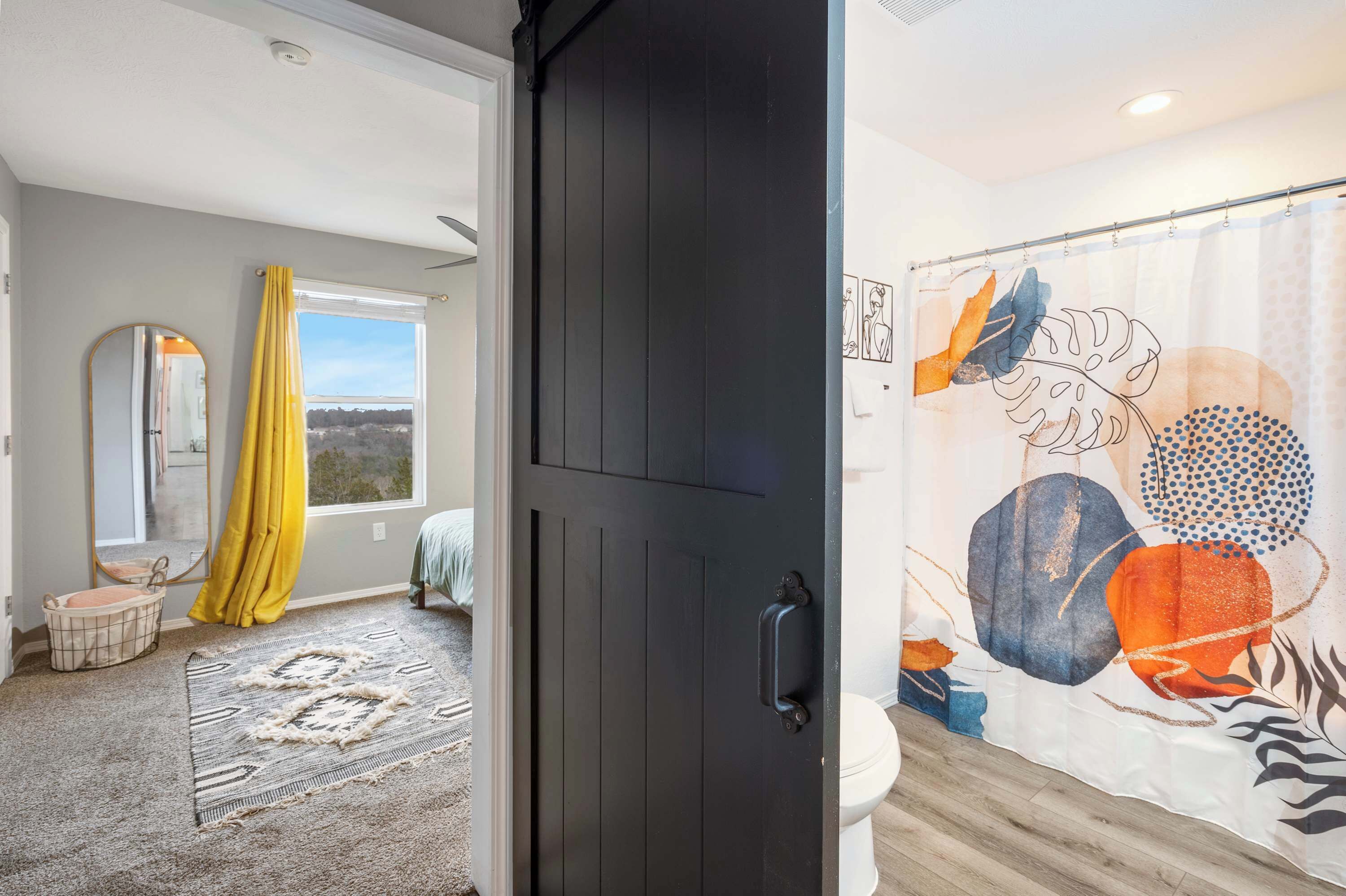 Une salle de bain avec un rideau de douche orné de motifs abstraits, des toilettes et une fenêtre donnant sur un paysage.