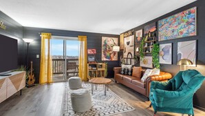 Ein modernes Wohnzimmer mit Sofa, Sessel, Couchtisch und Teppich. Es gibt ein Fenster mit Vorhängen, ein Bücherregal und eine Wand mit gerahmten Kunstwerken.