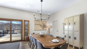 A dining area with a wooden table, grey chairs, a chandelier, and a view through large glass doors. - Big Bear Bungalow Stay on Ozark Waterfront (Gravois Mills)