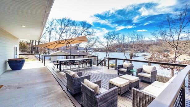 A covered outdoor deck with wicker furniture, a table, and chairs, overlooking a body of water and trees. - Big Bear Bungalow Stay on Ozark Waterfront (Gravois Mills)