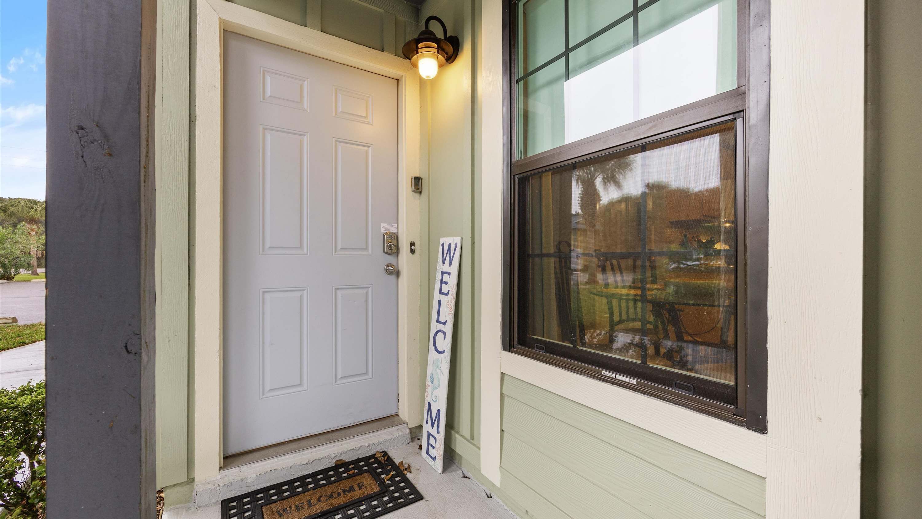 A white door with a 'WELCOME' sign, a window with a view of outdoor furniture, and a doorbell mat.