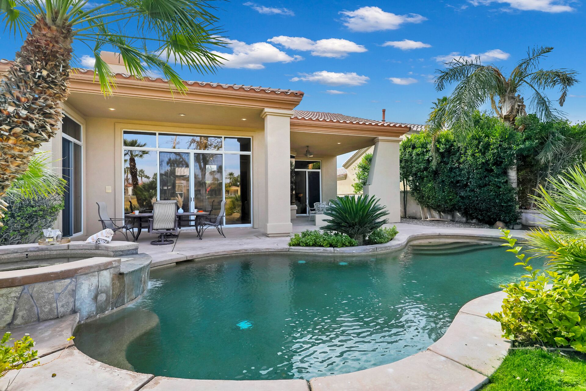 A pool with a curved edge, a patio with chairs and a table, and a covered area with a glass door.