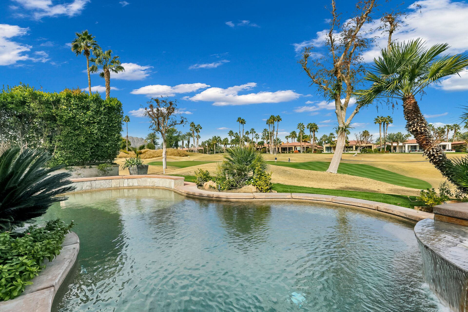 Ein Golfplatz mit Wasserspiel im Vordergrund, umgeben von Palmen und blauem Himmel.