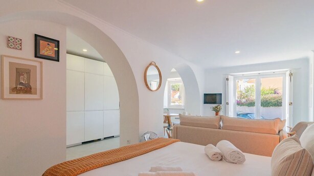 A modern hotel room with a large bed, a sitting area with a sofa, a dining table, and a view of the outdoors. - Chalet Maria da Assunção - Studio (Cascais)