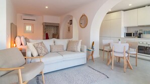 A modern living room with a white sofa, two armchairs, a small side table, and a kitchen area with white cabinets and appliances. - Chalet Maria da Assunção - Studio (Cascais)