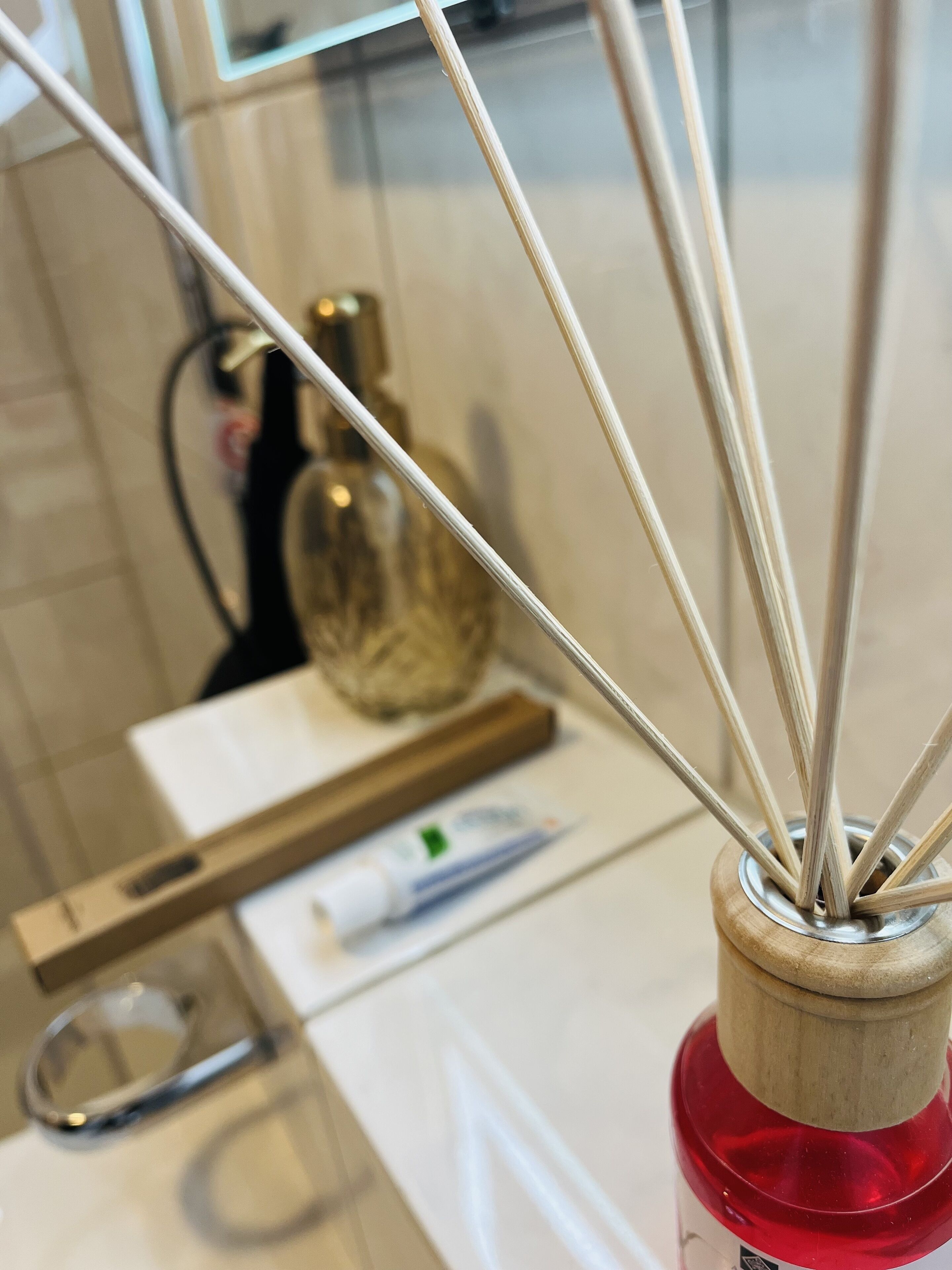 Detailaufnahme eines Holz-Reeds Diffusers mit mehreren Röhrchen in einem klaren Glasbehälter, platziert auf einer weißen Oberfläche mit rotem Sockel.