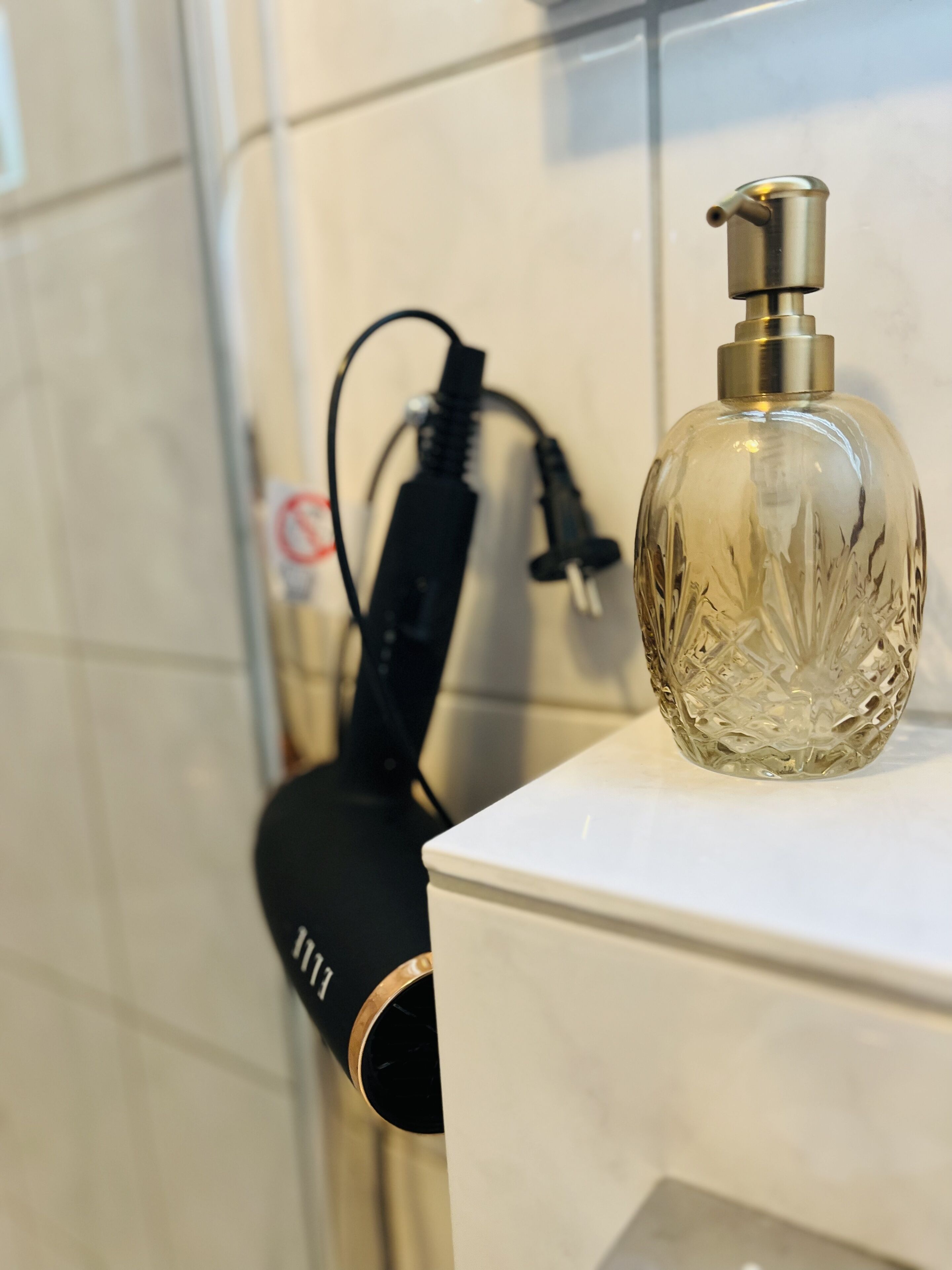 Een badkamer met een wit wastafelmeubel, een glazen fles met pompje en een zwarte haardroger op het aanrecht.