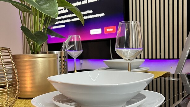 Un comedor con una mesa dispuesta para dos personas, una planta en maceta y un televisor que muestra texto al fondo.