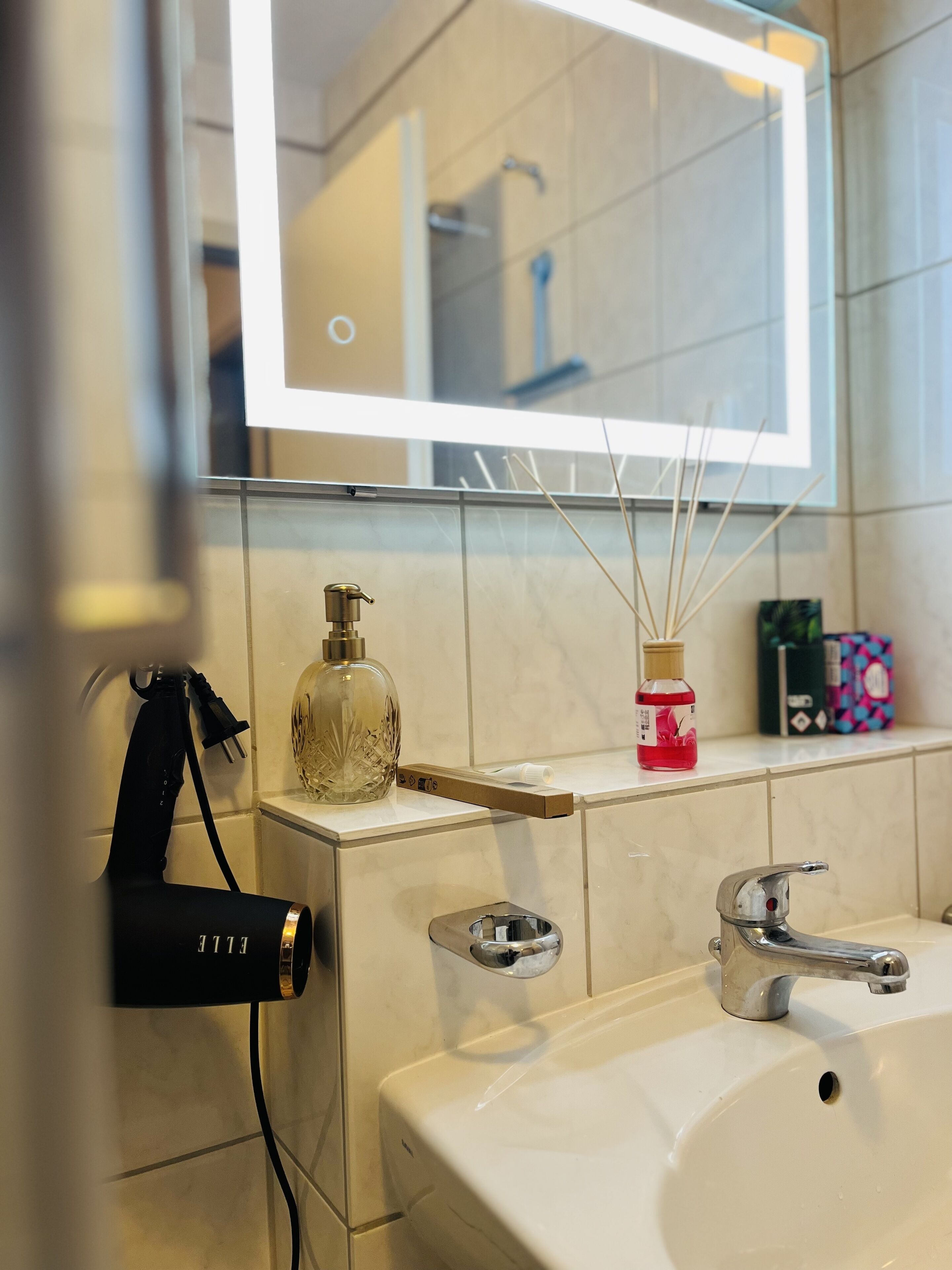 A bathroom with a mirror, a sink, a hairdryer, and a bottle of reed diffuser.