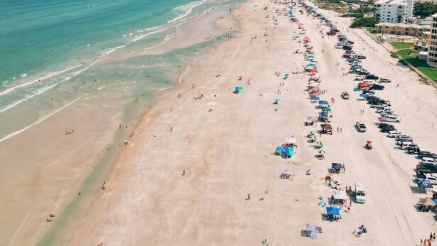 A beach with a long stretch of umbrellas and beachgoers, a parking lot, and buildings in the background.
