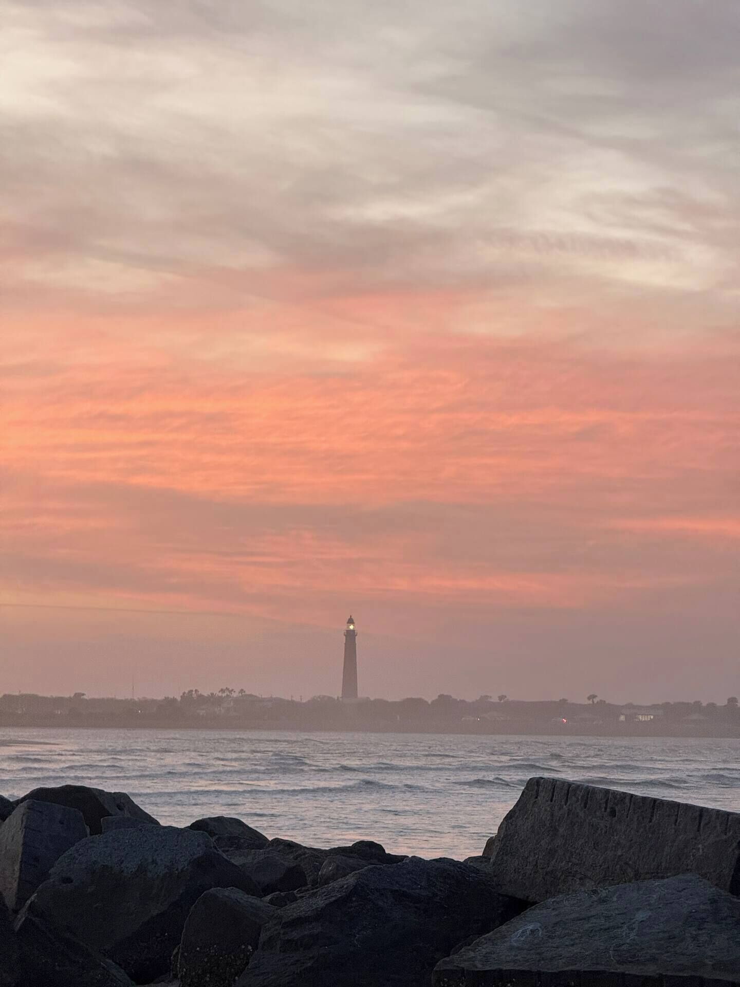 Um farol à beira-mar, com um primeiro plano rochoso e um céu ao pôr do sol.