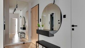 Un pasillo moderno con una puerta de madera, un espejo redondo, un banco negro y una pequeña mesa con plantas.
