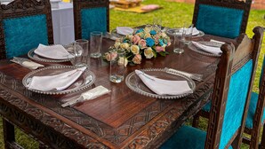 Una mesa de comedor de madera al aire libre, con sillas, un centro de mesa floral y un cielo despejado al fondo.