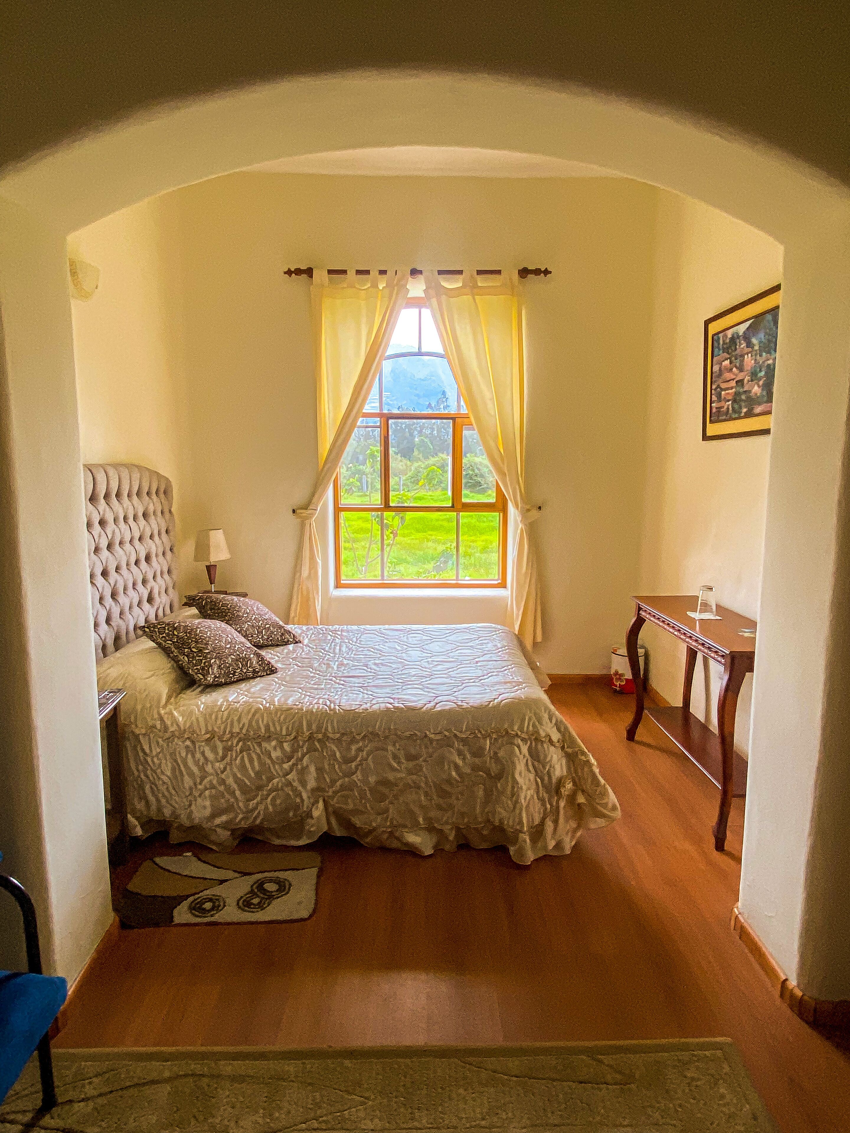 A bedroom with a large window, a bed with a headboard, a bedside table, and a small table with a glass.