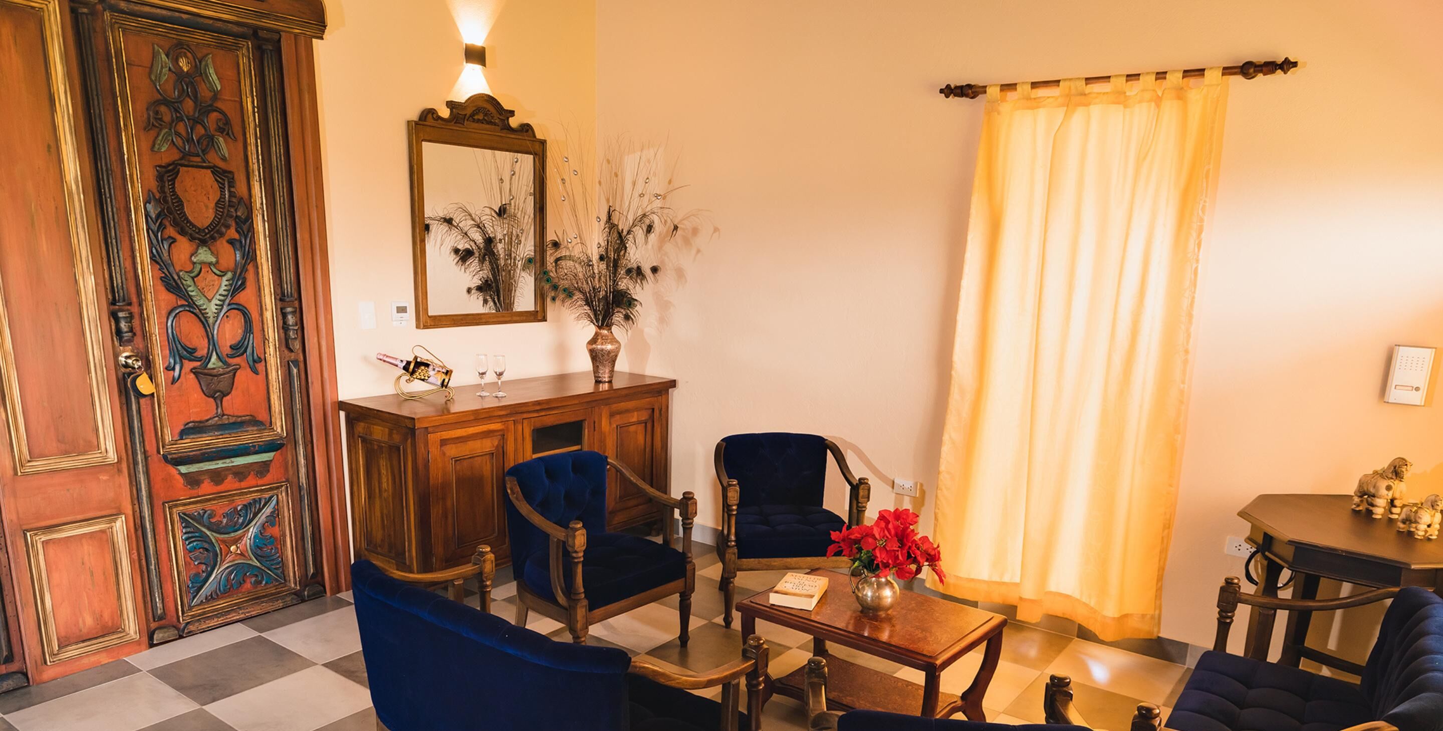 Una habitación con un armario de madera, un espejo, un jarrón con flores, una silla azul, una mesa y una cortina.