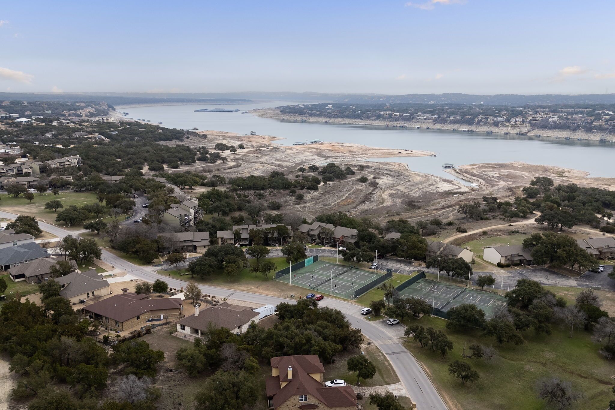 An aerial view of a residential area with houses, a tennis court, and a large body of water in the background.