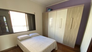 A bedroom with a bed, wooden floor, wardrobe, and a window with blinds. - Casa do Bert Itanhaén (Itanhaém)
