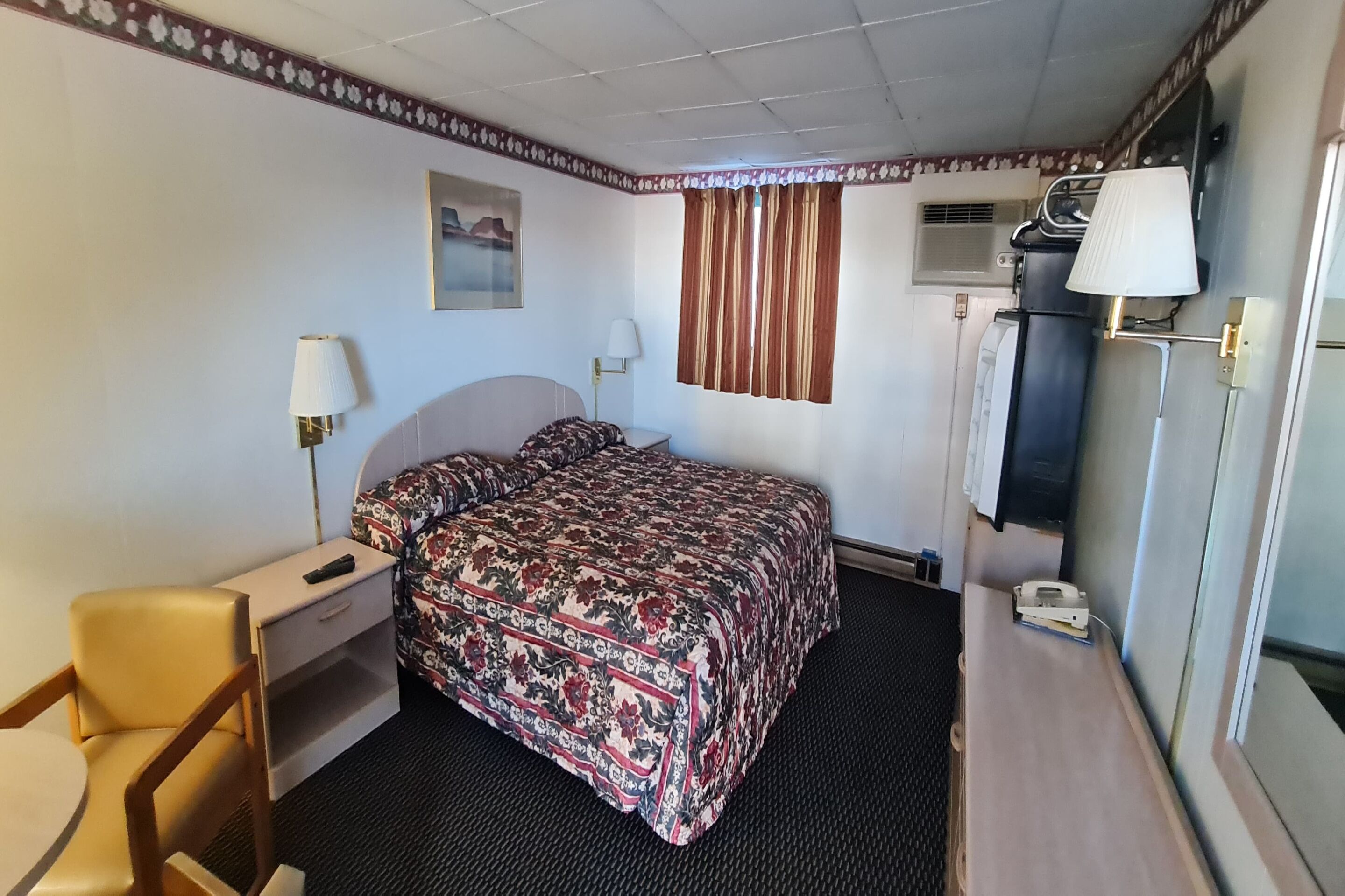 A hotel room with a bed, a desk, a chair, a lamp, and a window with curtains.
