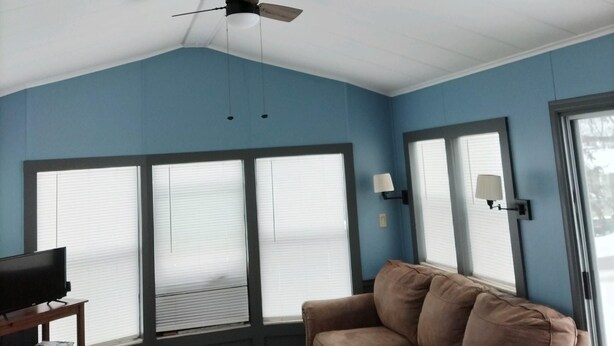 A room with a ceiling fan, large windows with blinds, a brown sofa, a TV on a wooden stand, and a lamp on the wall.