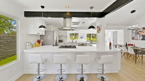 A modern kitchen with a large island, white bar stools, and a dining area with a table and chairs. - Luxury Villa: Pool, Spa, Golf Views  Sleeps 12 (Indio)