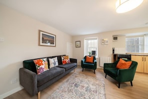 A living room with a sofa, two armchairs, a rug, and a kitchen area with a stove.