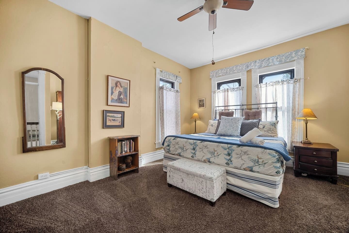A bedroom with a bed, a ceiling fan, a nightstand, a bedside table, a lamp, a bookshelf, and a window with curtains.