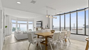 Un comedor moderno con una mesa de madera y sillas blancas, ventanas amplias con vista al mar, y un sofá blanco al fondo.