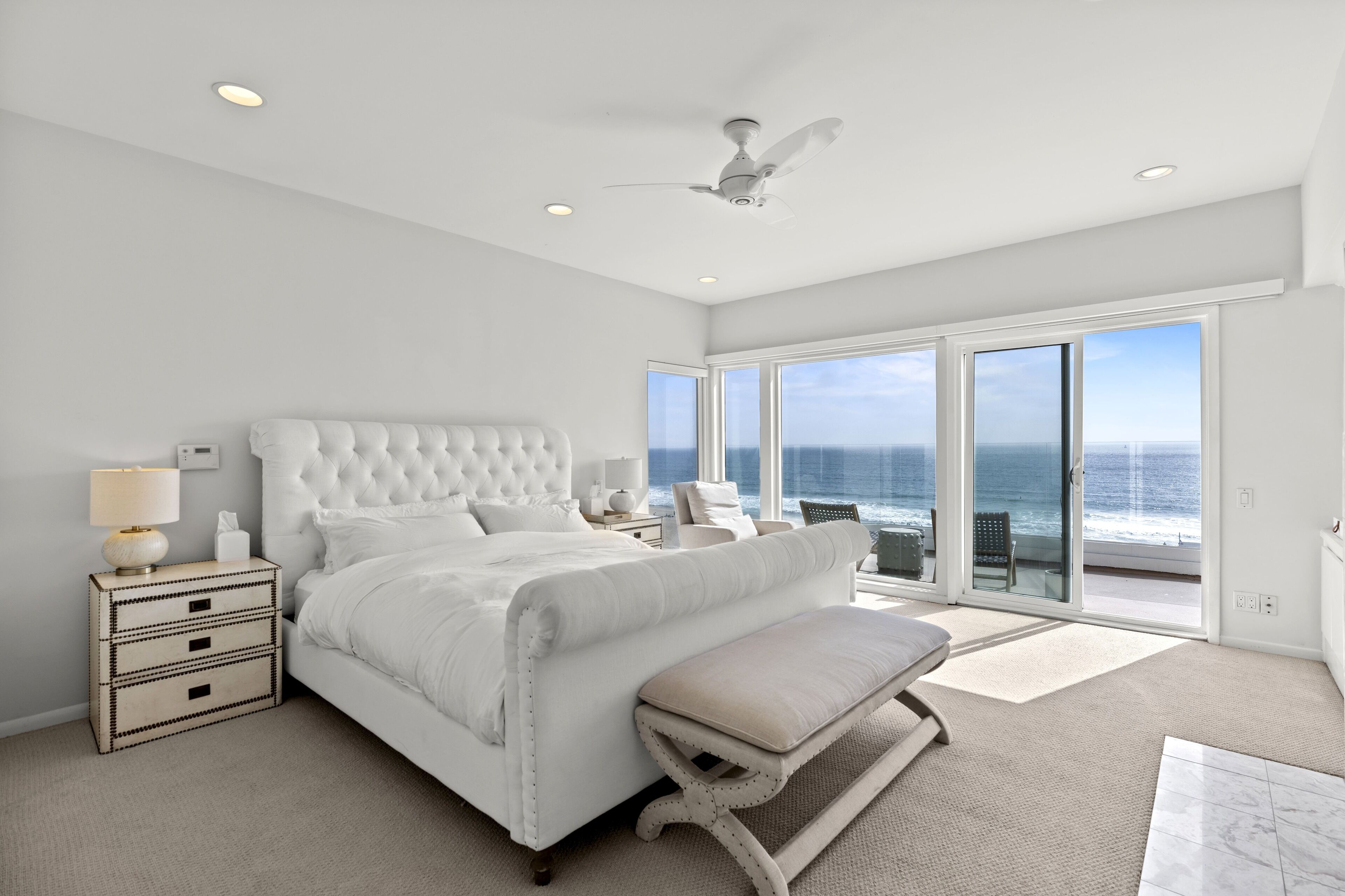 A spacious bedroom with a large bed, a ceiling fan, and a view of the ocean through floor-to-ceiling windows.