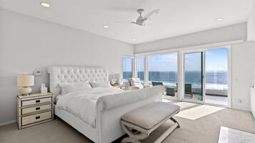 A spacious bedroom with a large bed, a ceiling fan, and a view of the ocean through floor-to-ceiling windows.