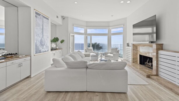 Una sala moderna con un sofá blanco modular, una chimenea y un ventanal con vistas al océano.