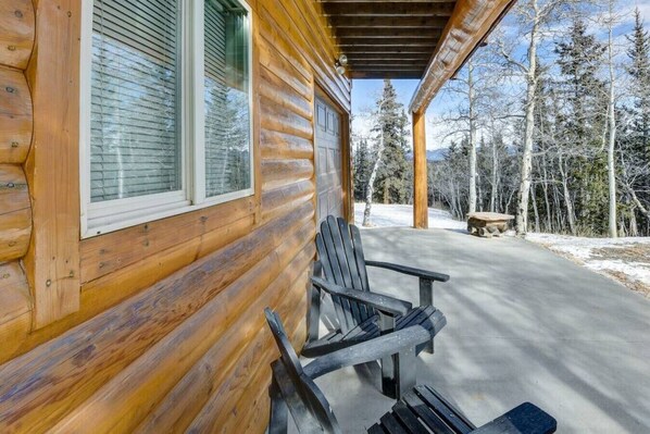 A wooden cabin with a porch, two Adirondack chairs, and a stone bench.