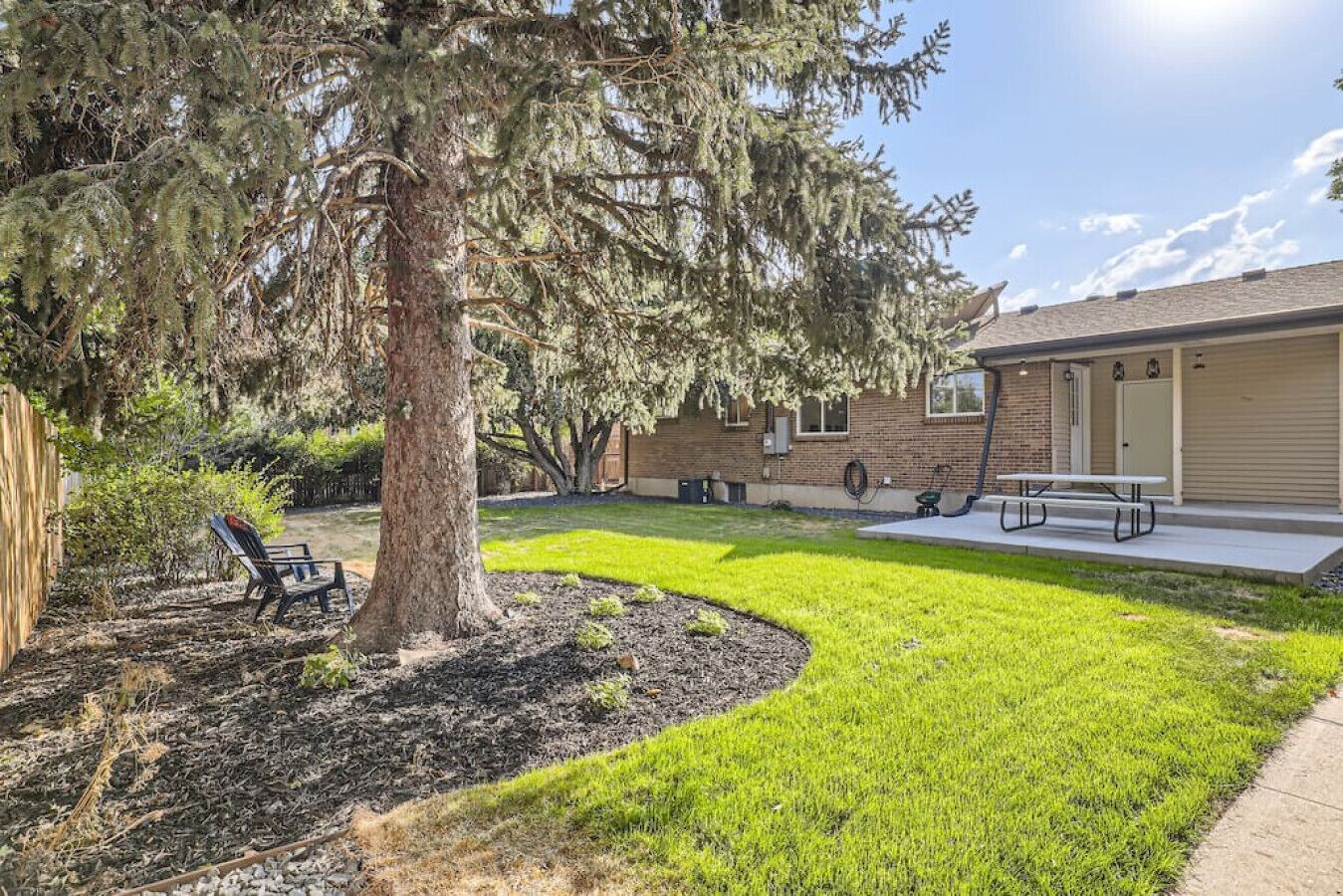 A residential backyard with a well-maintained lawn, a large tree, a picnic table, and a house with a brick exterior.