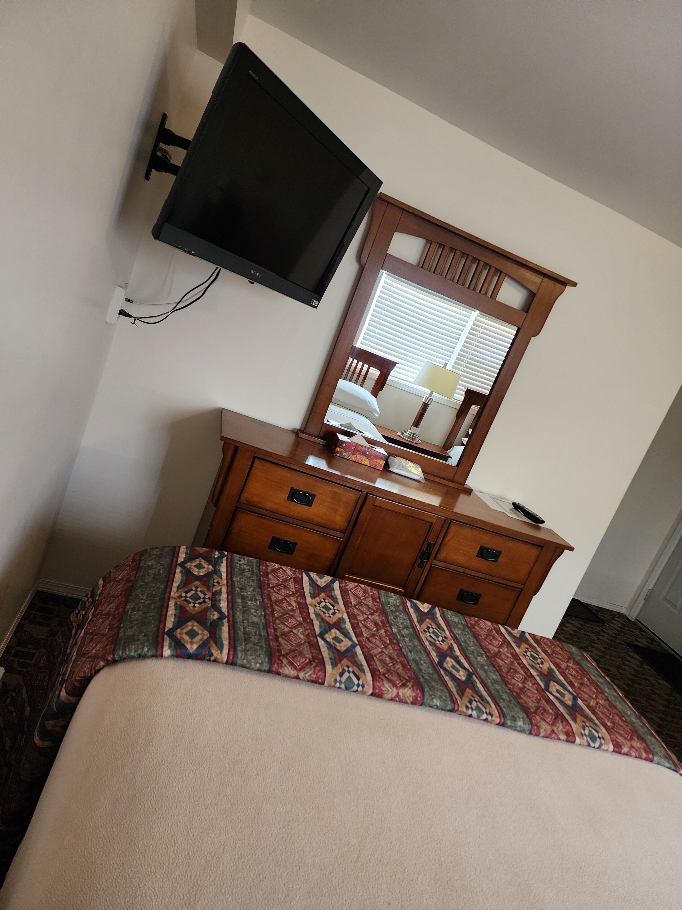 A hotel room with a bed, a wooden dresser, a flat-screen TV mounted on the wall, and a window with blinds.