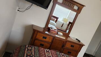 A hotel room with a bed, a wooden dresser, a flat-screen TV mounted on the wall, and a window with blinds.