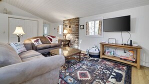 A cozy living room with a couch, coffee table, and a patterned rug. There's a stone wall, a television, and a wooden shelf with items on it. - Family Friendly Cabin I Firepit+Hot Tub I Poconos (Pocono Pines)