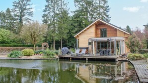 Eine Holzblockhütte mit überdachter Veranda, einem Teich davor und umliegenden Bäumen.