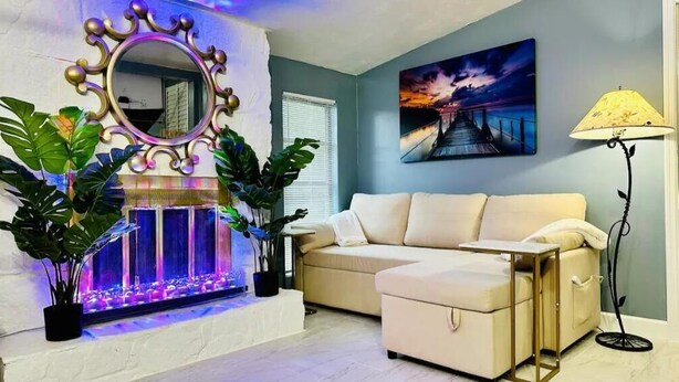 A living room with a beige sofa, a decorative mirror, a painting of a sunset, and a floor lamp.
