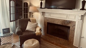 A living room with a fireplace, a television mounted above it, a comfortable armchair, and a small round ottoman. - Beautiful home on Lake Hamilton  ! (Lake Hamilton)