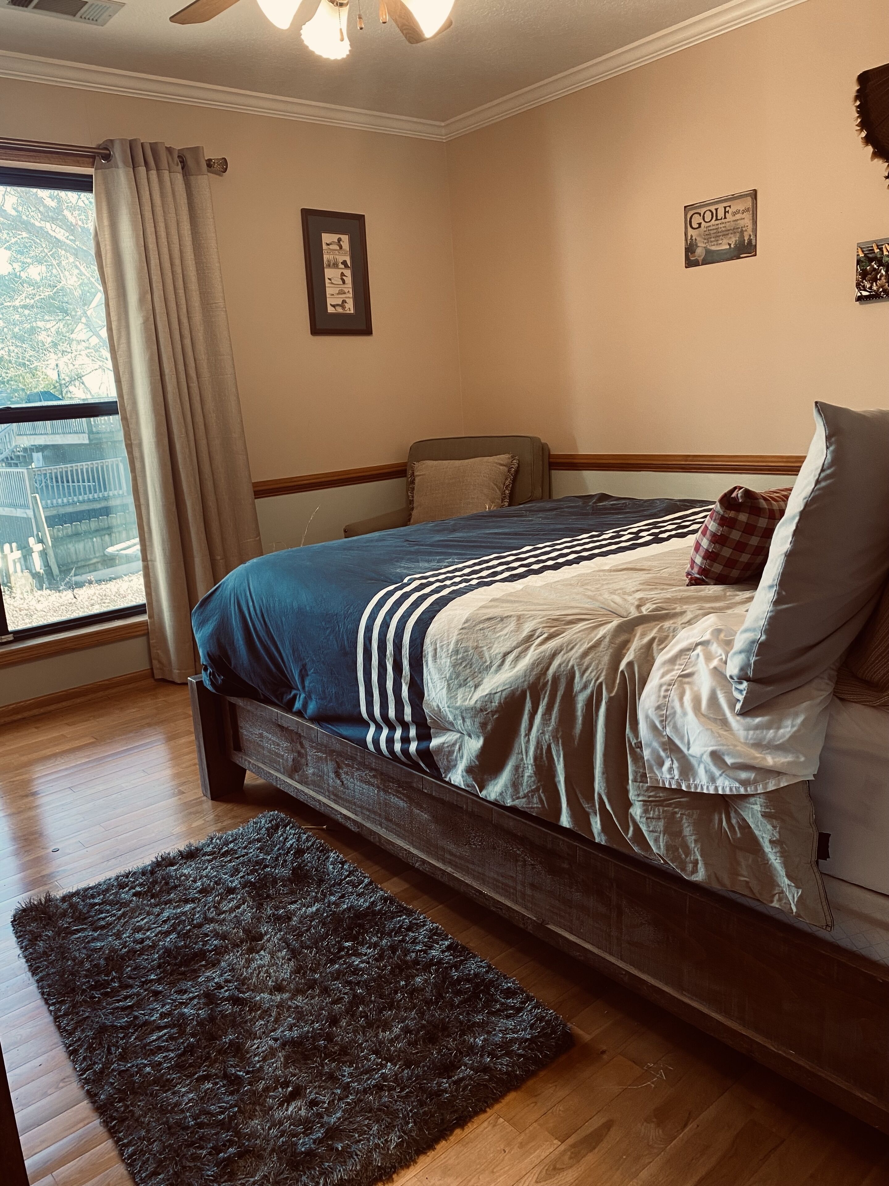 Un dormitorio con una cama de madera, una alfombra oscura, una ventana con cortinas y un ventilador de techo.