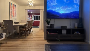 Ein modernes Wohnzimmer mit Flachbildfernseher, Holzboden, einer Essecke mit Stühlen, einem Tisch und einem Bücherregal mit Büchern und Dekoration.